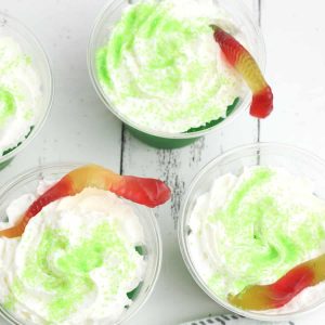 Green jello shots topped with whipped cream and gummy worms on a white table.
