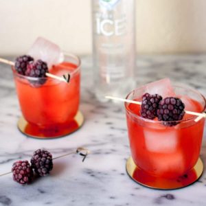 Two berry cocktails with blackberries on cocktail picks on a marble table with a bottle of sparkling ice.