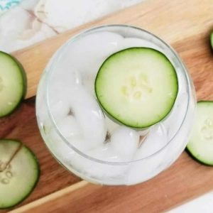 A vodka cocktail garnished with a fresh slice of cucumber on a cutting board.
