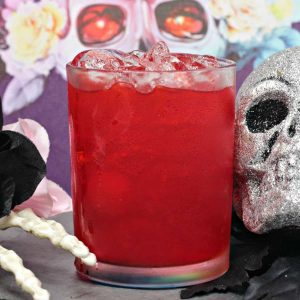 A silver glitter skull sits next to a spiced wine cocktail in front of a Halloween backdrop and a bony skeleton hand.