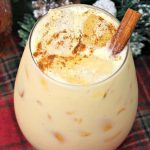 A glass of boozy eggnog with a cinnamon stick on a table decorated for Christmas.