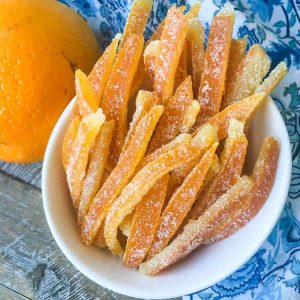 A bowl of candied orange peels on a blue and white napkin next to an orange.
