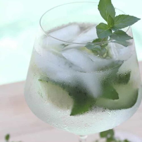 A wine glass filled with ice water, cucumber slices, and fresh mint next to a swimming pool.