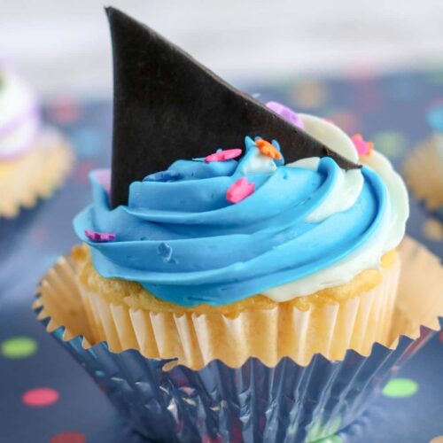 A vanilla cupcake with blue and white swirled frosting with a black shark fin on top.