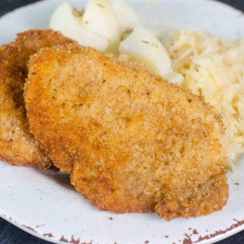 Two pieces of pork schnitzel on a white plate with a side of sauerkraut and potatoes.